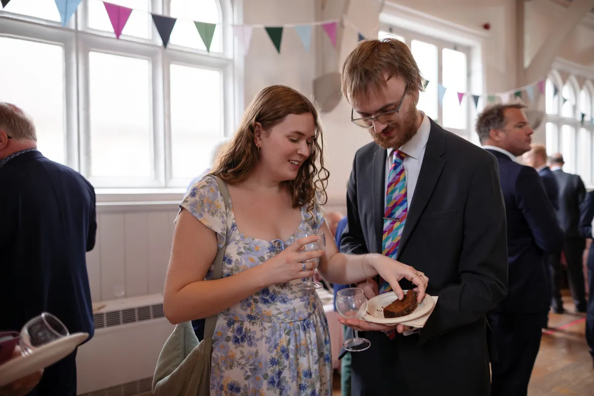 18_TJP_2025_08_30_0456 A woman in a floral dress and a man in a suit with a colourful tie stand together at a social event, smiling and holding cake and drinks. Candid moments like these are expertly captured by an Octagon Barn Norwich Wedding Photographer.