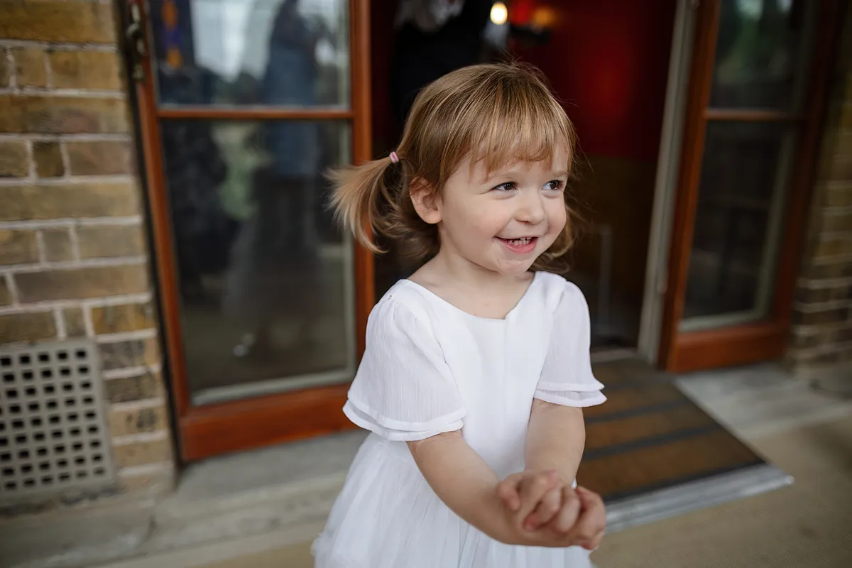 19_TJP_2025_08_16_0274 A young girl with light brown hair in plaits, wearing a white dress, smiles and clasps her hands whilst standing in front of an open doorway—perfect for a charming Pitzhanger Manor Wedding Photography moment.