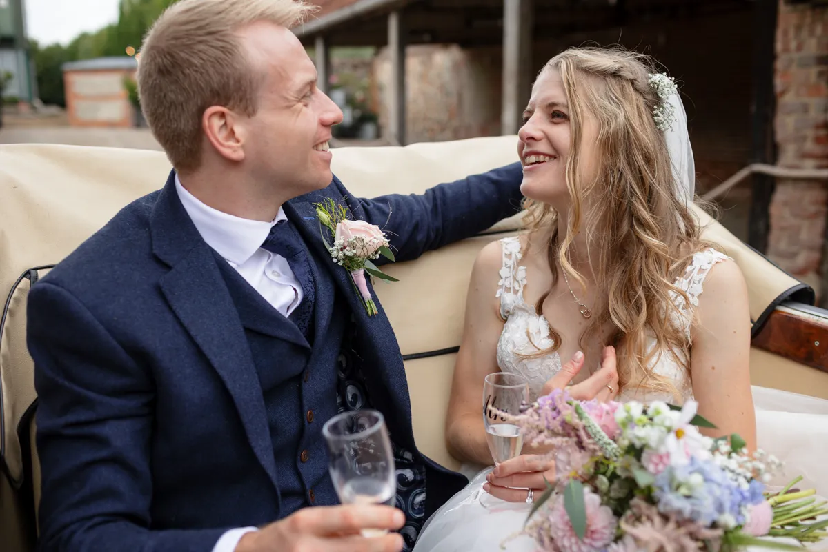 19_TJP_2025_08_30_0501 A bride and groom sit close together, smiling and holding glasses of champagne. Captured by Octagon Barn Norwich Wedding Photographer, she holds a bouquet in her lace-detailed dress while he wears a navy suit with a buttonhole.