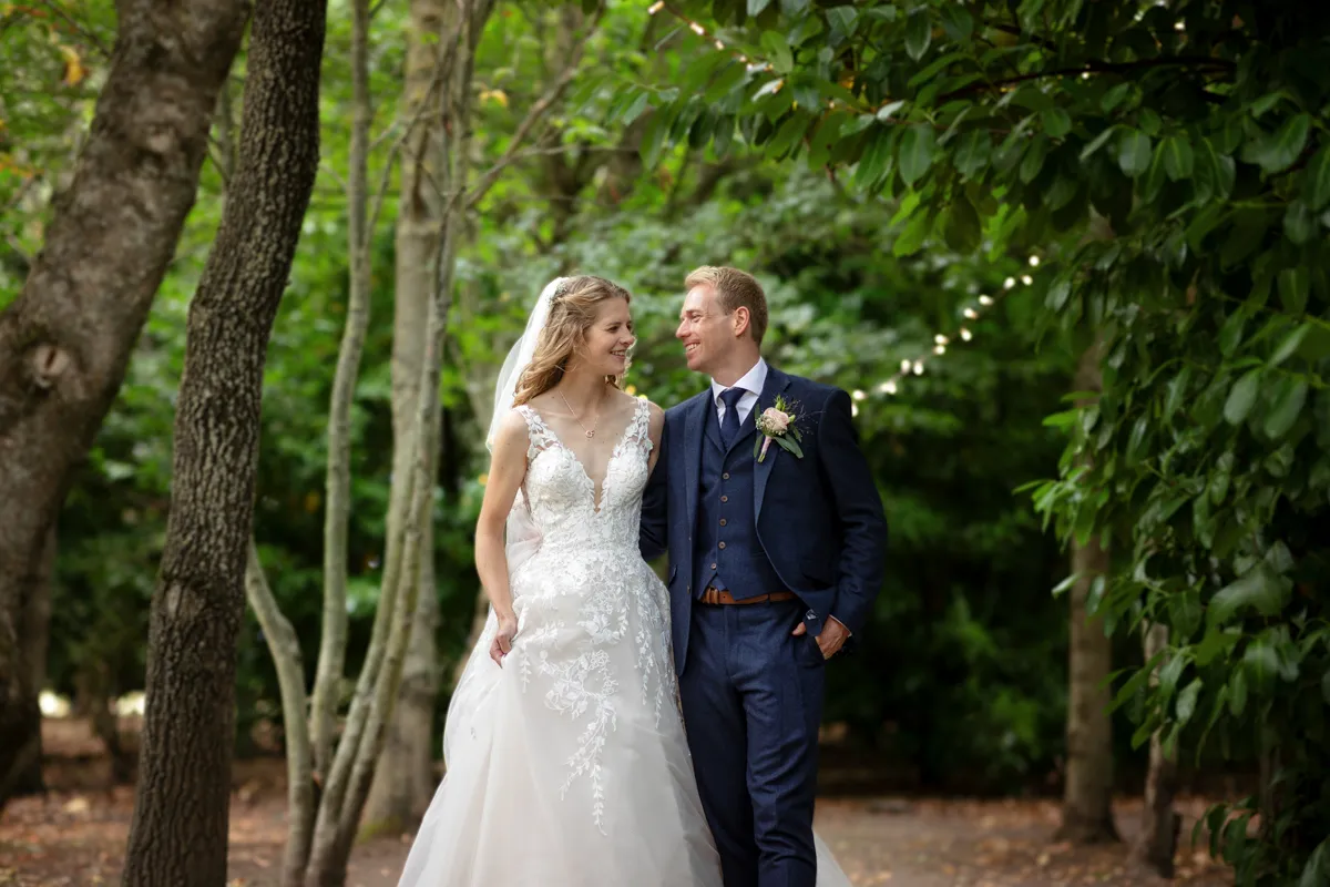 20_TJP_2025_08_30_0528 A bride in a white wedding dress and a groom in a navy suit walk together outside, smiling at each other, surrounded by green trees and soft fairy lights captured beautifully by Octagon Barn Norwich Wedding Photographer.