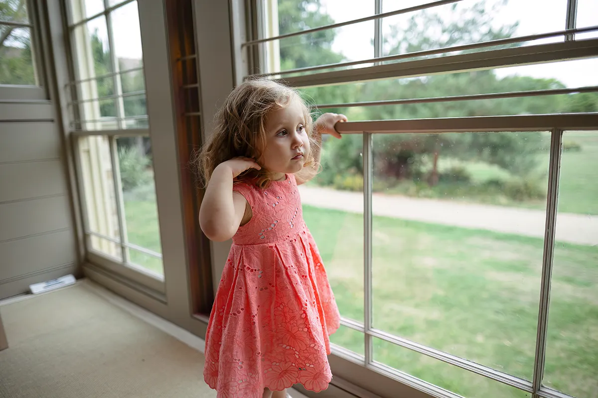 21_TJP_2025_08_16_0277 A young girl in a coral dress stands by large windows at Pitzhanger Manor, looking outside at a green lawn and trees with a thoughtful expression—capturing a serene moment of wedding photography.