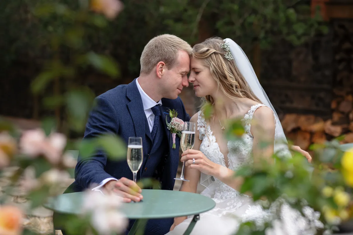 21_TJP_2025_08_30_0571 A bride and groom sit closely at a small outdoor table, touching foreheads and smiling with their eyes closed, holding champagne flutes, surrounded by greenery and flowers—captured beautifully by an Octagon Barn Norwich Wedding Photographer.