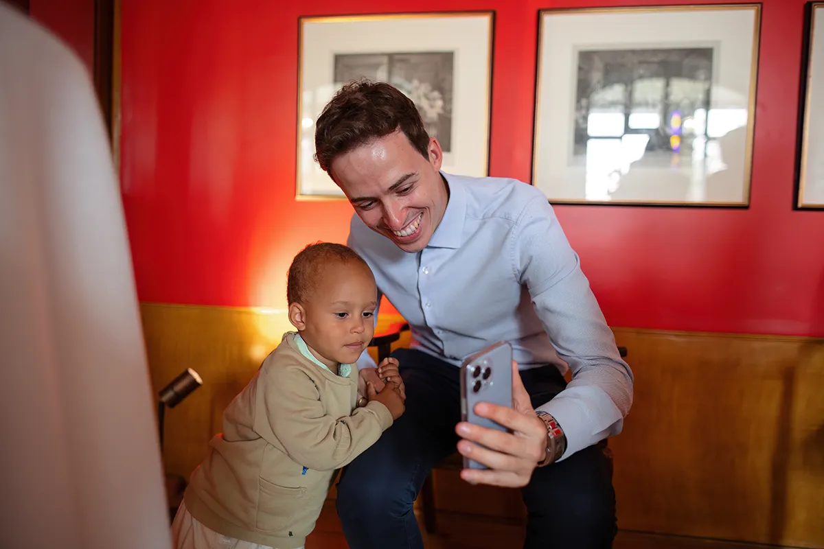 22_TJP_2025_08_16_0279 A smiling man in a light blue shirt holds a mobile, taking a selfie with a young child in a beige jacket. Indoors, they stand before a bright red wall with framed pictures—a candid moment reminiscent of Pitzhanger Manor Wedding Photography.
