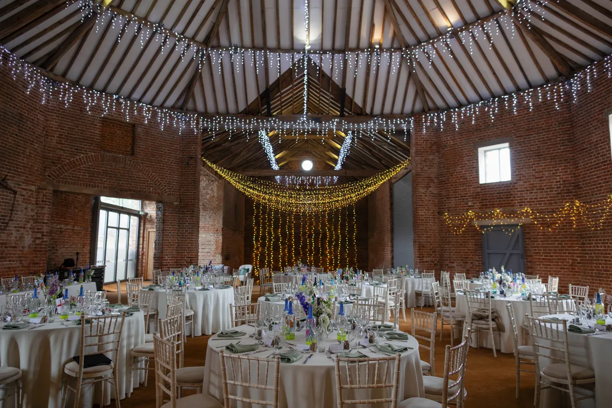 22_TJP_2025_08_30_0585 A rustic wedding reception venue with round tables and white chairs, decorated with floral centrepieces. Fairy lights hang from the beamed ceiling, perfect for an Octagon Barn Norwich Wedding Photographer to capture the warm, festive atmosphere.