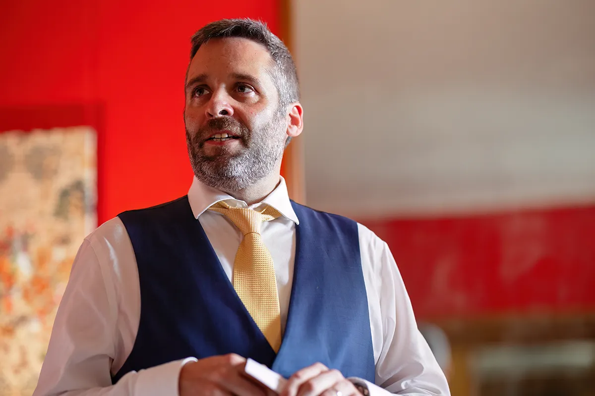 23_TJP_2025_08_16_0338 A man with short brown hair and a beard, dressed in a white shirt, yellow tie, and dark blue waistcoat, stands indoors before a red wall, looking slightly to the side and smiling—captured in elegant Pitzhanger Manor Wedding Photography style.