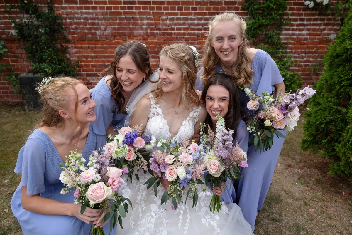 23_TJP_2025_08_30_0615 A bride in a white dress sits smiling, surrounded by four bridesmaids in light blue dresses, all holding colourful bouquets, posing happily outdoors in front of a brick wall and greenery by Octagon Barn Norwich Wedding Photographer.
