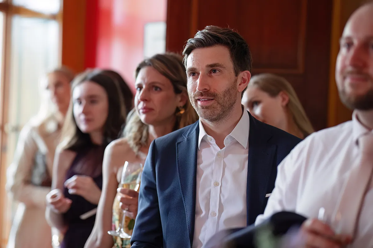 24_TJP_2025_08_16_0342 A group of people dressed in formal attire stand indoors at a Pitzhanger Manor Wedding Photography session, attentively watching something off-camera. The man in front wears a navy suit and white shirt, while a woman behind him holds a glass of champagne.