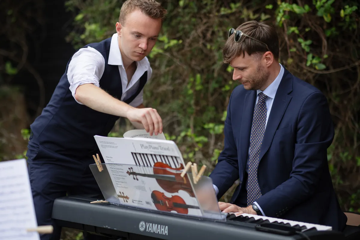 24_TJP_2025_08_30_0640 Two men, one in a suit playing a Yamaha keyboard and another adjusting sheet music by greenery, prepare near illustrations of piano and violin—captured perfectly by an Octagon Barn Norwich Wedding Photographer.
