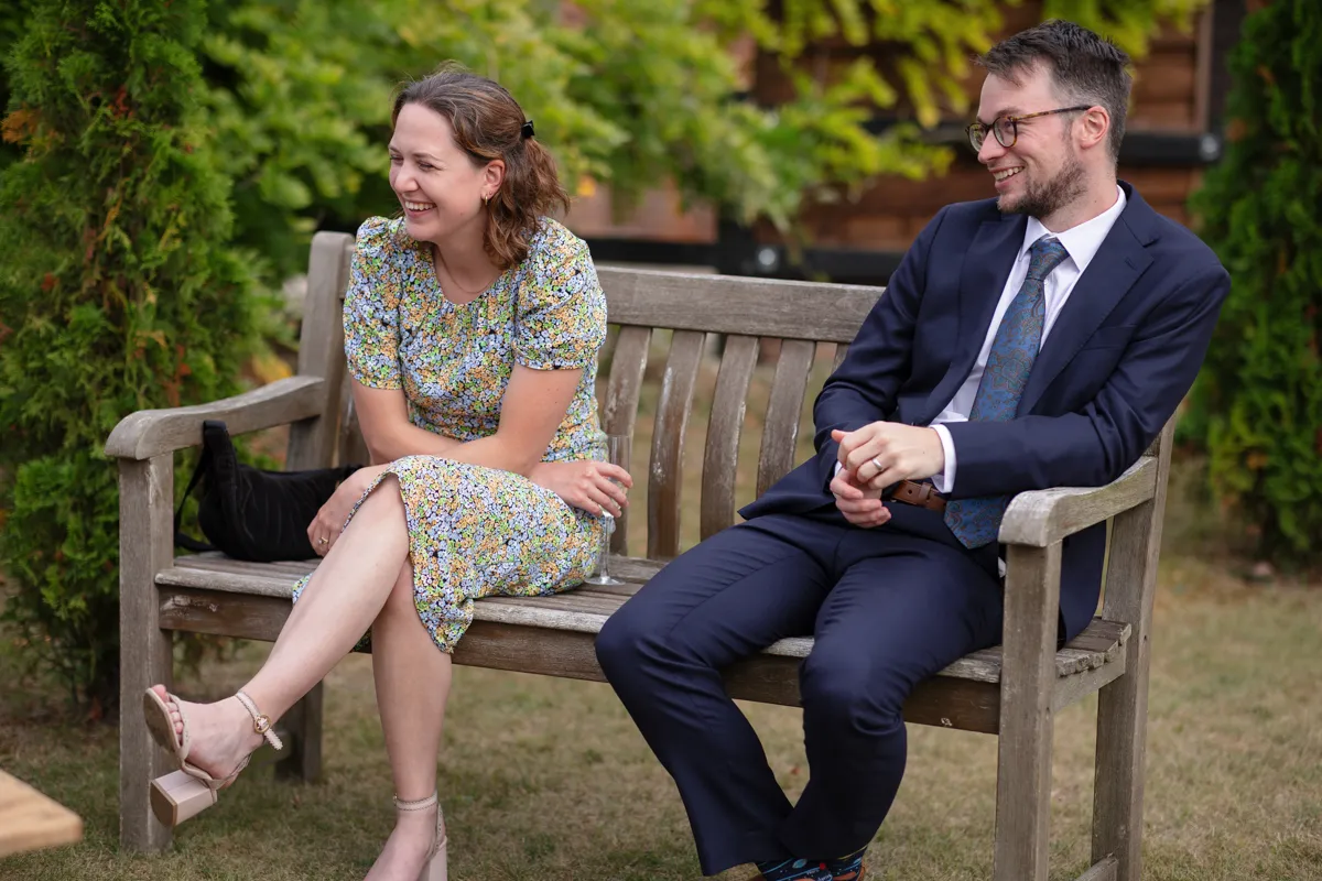25_TJP_2025_08_30_0655 A woman in a floral dress and a man in a suit sit on a wooden bench outdoors, both smiling and looking to the side. Greenery and grass are visible in the background, captured beautifully by Octagon Barn Norwich Wedding Photographer.
