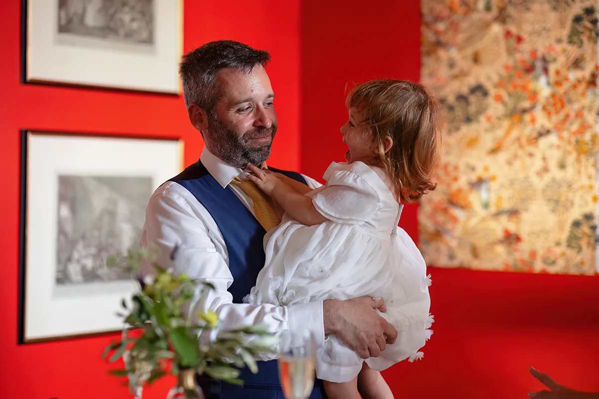 26_TJP_2025_08_16_0384 A man in a blue waistcoat and yellow tie smiles whilst holding a young girl in a white dress. They are indoors, with red walls adorned with art and flowers—a joyful moment of Pitzhanger Manor Wedding Photography.