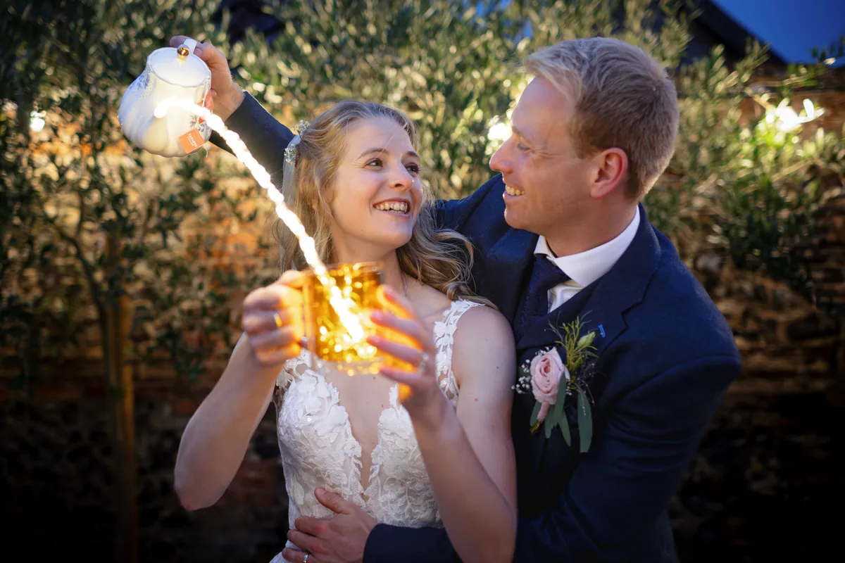 26_TJP_2025_08_30_0896 A smiling bride in a white dress holds a glowing mug as the groom, in a dark suit, pours liquid from a teapot. Captured by Octagon Barn Norwich Wedding Photographer, they embrace outdoors at night, illuminated by warm lights, looking joyfully at each other.