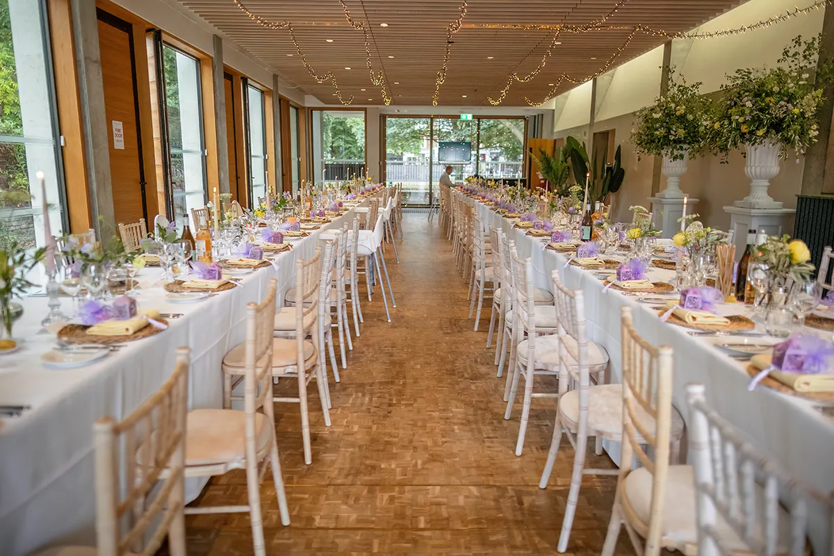 27_TJP_2025_08_16_0390 A long, elegantly decorated wedding reception hall with white-covered tables, floral centrepieces, purple accents, and fairy lights. Perfect for Pitzhanger Manor Wedding Photography, large windows line one side of the room.
