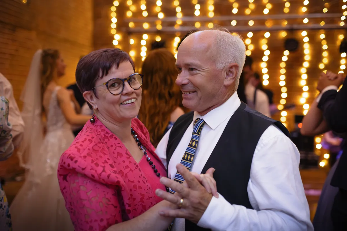 27_TJP_2025_08_30_0920 An older couple smiles while dancing together at a festive indoor event with fairy lights in the background. Other guests, including a bride in white, join the celebration—captured beautifully by an Octagon Barn Norwich Wedding Photographer.