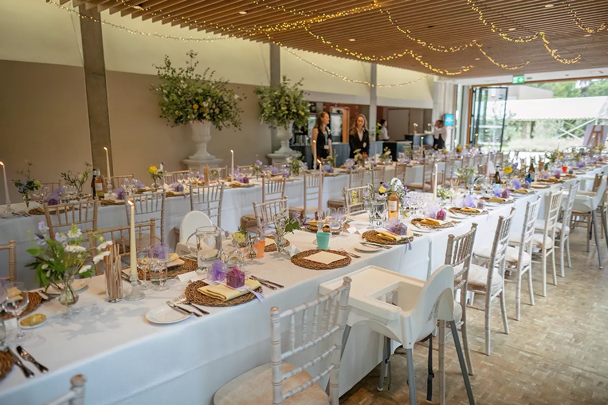 28_TJP_2025_08_16_0392 A wedding or event reception hall with long tables set for a meal, adorned with flowers, candles, and fairy lights. Elegant chairs and highchairs surround the tables as guests mingle—perfect for capturing Pitzhanger Manor Wedding Photography moments.