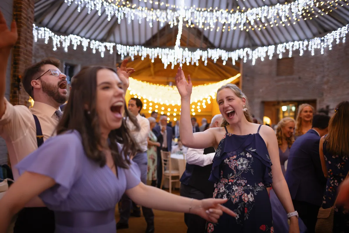 28_TJP_2025_08_30_0930 A group of people joyfully dancing and laughing at a festive indoor event, with twinkling fairy lights overhead, creating a warm and lively atmosphere—captured beautifully by Octagon Barn Norwich Wedding Photographer.