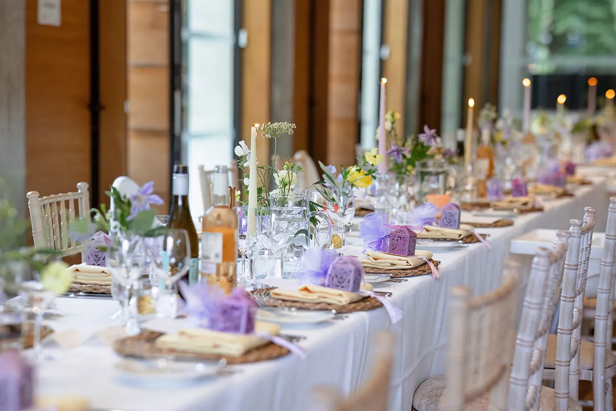 29_TJP_2025_08_16_0403 A long, elegant dining table set for an event—perfect for Pitzhanger Manor Wedding Photography—adorned with flowers, candles, wine bottles, glassware, and purple gift boxes on white tablecloths, surrounded by light wooden chairs.