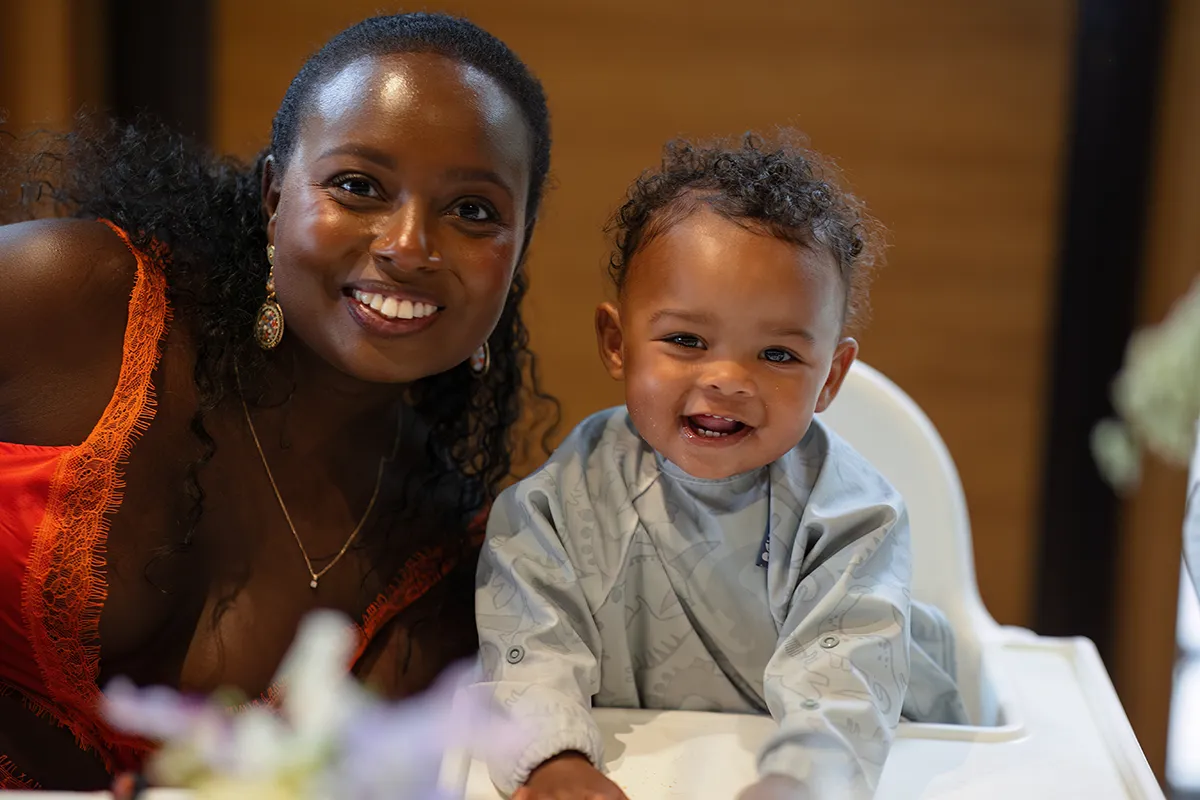 30_TJP_2025_08_16_0430 A smiling woman in an orange dress leans next to a cheerful baby with curly hair in a highchair, both looking at the camera. The softly blurred background adds warmth, evoking the candid charm of Pitzhanger Manor Wedding Photography.