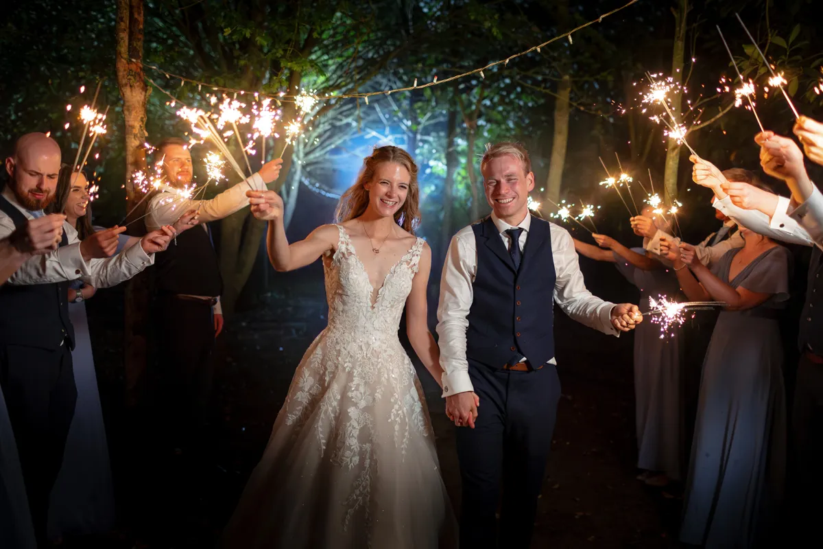 30_TJP_2025_08_30_1027 A bride and groom walk hand-in-hand at night, smiling, surrounded by guests holding sparklers under twinkling fairy lights—captured perfectly by an Octagon Barn Norwich Wedding Photographer for a joyful, festive atmosphere.