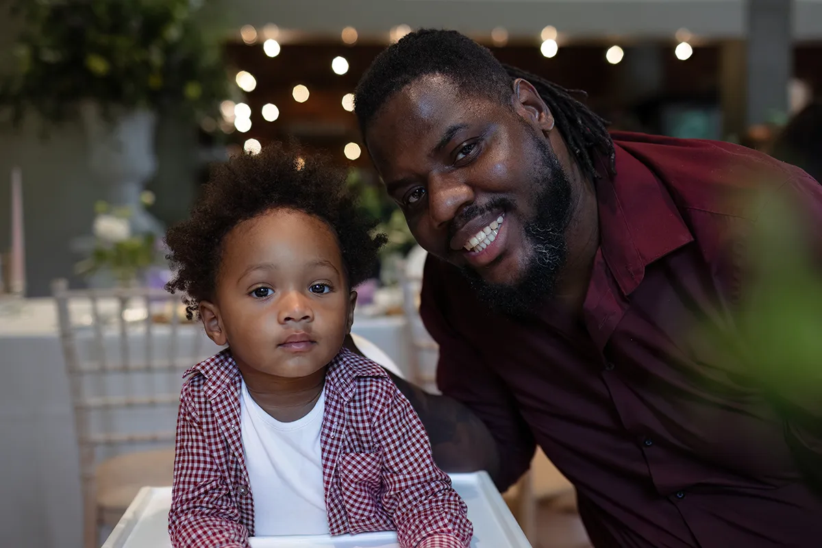 31_TJP_2025_08_16_0477 A smiling man in a maroon shirt leans next to a young child with curly hair and a checked shirt, captured indoors at a decorated event. Blurred lights and tables in the background evoke the charm of Pitzhanger Manor Wedding Photography.