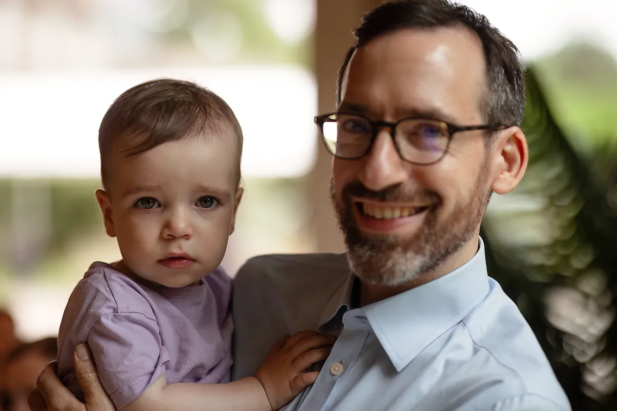 32_TJP_2025_08_16_0485 A man with glasses smiles whilst holding a young child in his arms. Perfect for Pitzhanger Manor Wedding Photography, the child wears a light purple shirt and looks at the camera with a neutral expression; the softly blurred background adds warmth.