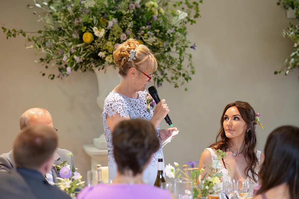 33_TJP_2025_08_16_0500 A woman in a light purple lace dress speaks into a microphone, holding a note, while a smiling bride in white looks on during a Pitzhanger Manor Wedding Photography session, surrounded by elegant flowers and greenery.