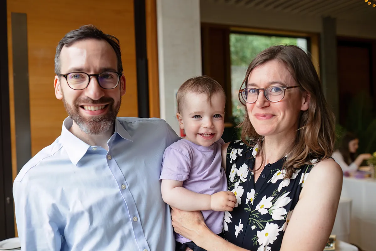 34_TJP_2025_08_16_0536 A man and woman wearing glasses smile at the camera, standing indoors. The woman holds a smiling toddler in her arms. This happy, close family radiates joy—captured with the warmth typical of Pitzhanger Manor Wedding Photography.