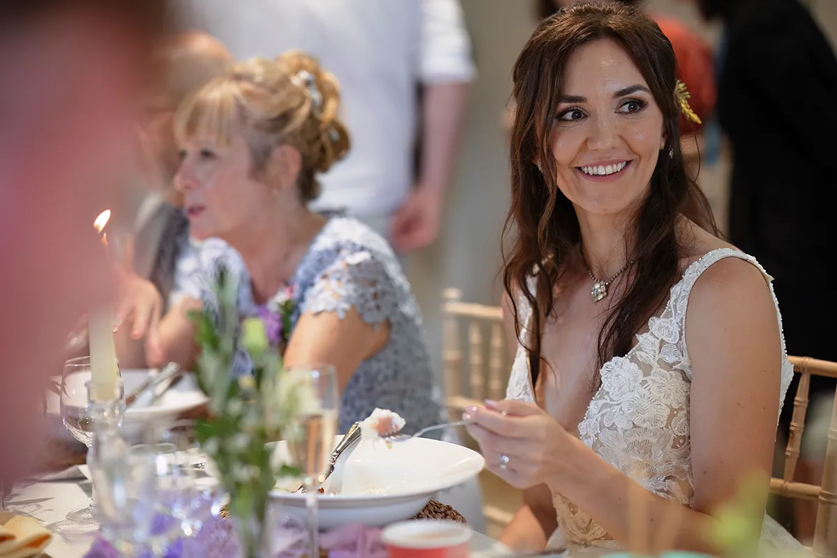 35_TJP_2025_08_16_0559 A smiling woman in a white lace dress sits at a decorated table with a bowl of food, holding a spoon. An older woman is beside her, with flowers and glasses on the table—capturing a joyful moment of Pitzhanger Manor Wedding Photography.