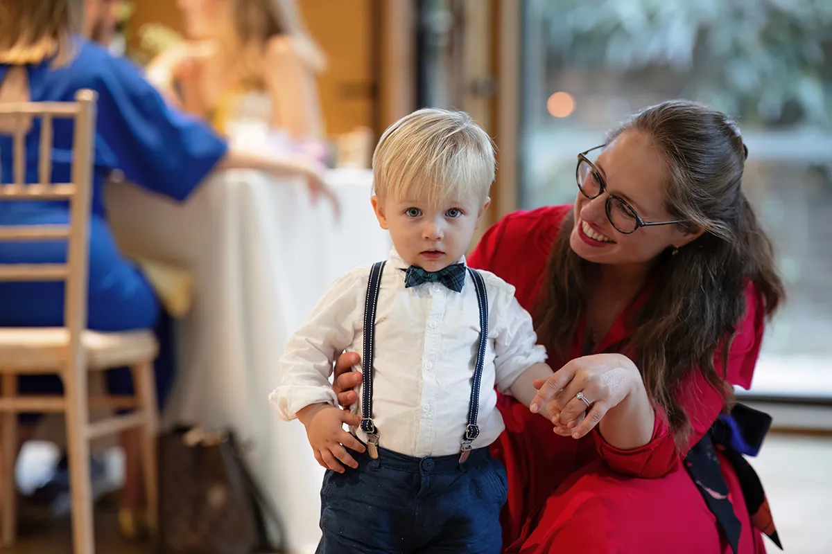 36_TJP_2025_08_16_0562 A young boy in a white shirt, bow tie, and braces stands while a smiling woman in glasses and a red dress holds his hand. Captured indoors with people and a table blurred behind, this tender moment reflects Pitzhanger Manor Wedding Photography.