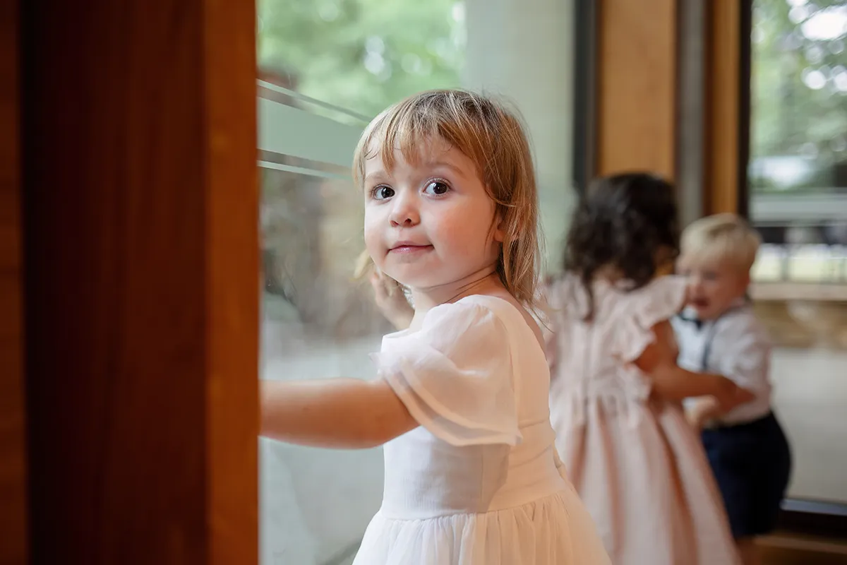37_TJP_2025_08_16_0569 A young girl in a white dress looks at the camera whilst standing by a window. In the background, two other children interact with each other in a softly lit indoor space—capturing the charm of Pitzhanger Manor Wedding Photography.