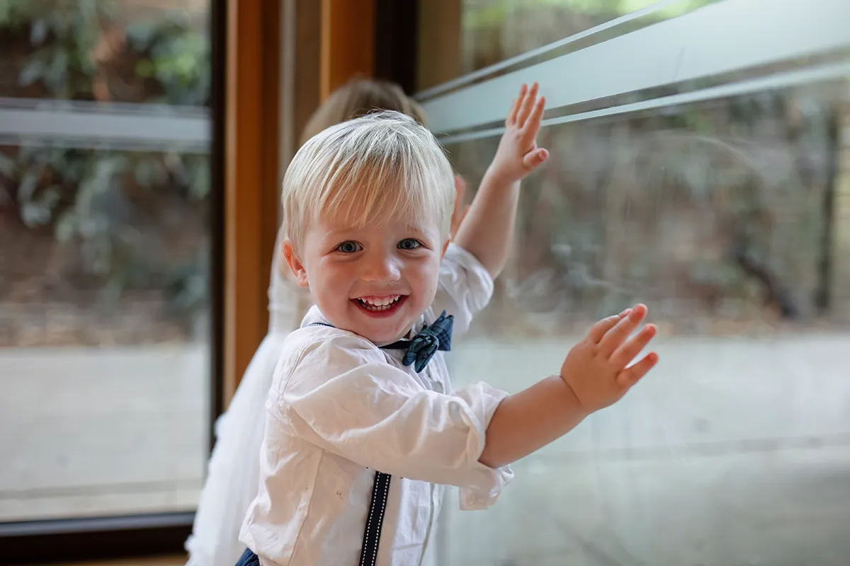 38_TJP_2025_08_16_0577 A young child with blonde hair, wearing a white shirt and blue braces, smiles and touches a glass window indoors—a candid moment captured in the style of Pitzhanger Manor Wedding Photography. Another child is partially visible in the background.