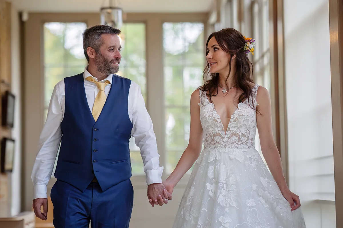 39_TJP_2025_08_16_0643 A bride in a white lace dress and a groom in a blue suit with a yellow tie walk hand in hand indoors, smiling warmly at each other—a candid moment captured with Pitzhanger Manor wedding photography.