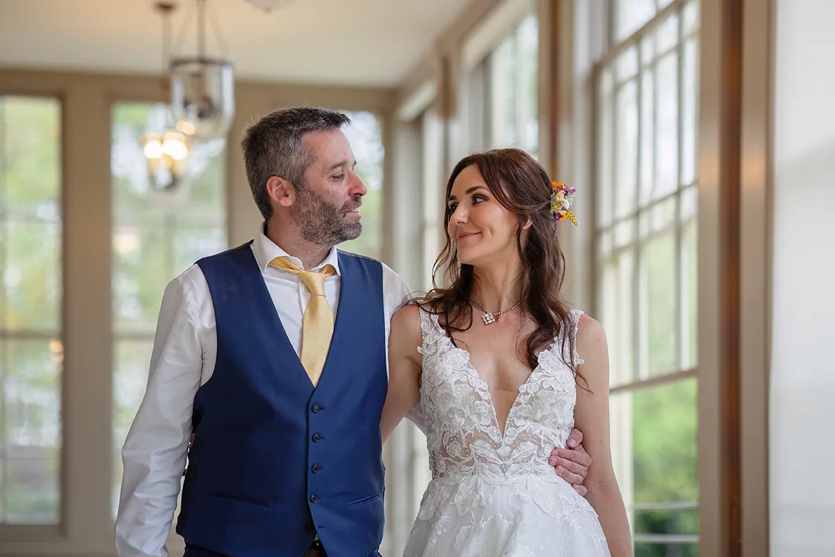 40_TJP_2025_08_16_0651 A bride and groom lovingly look at each other indoors in this Pitzhanger Manor Wedding Photography moment. The groom wears a blue waistcoat and yellow tie; the bride’s white lace dress and floral hairpiece glow in the natural light from large windows.