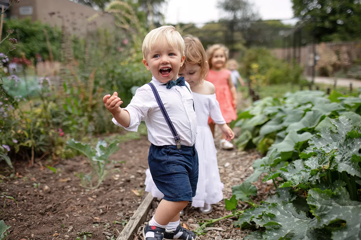 41_TJP_2025_08_16_0672 A young boy in braces and shorts smiles and walks on a garden path at Pitzhanger Manor, followed by other children, with green plants and soil beds around them—perfect for candid wedding photography moments.
