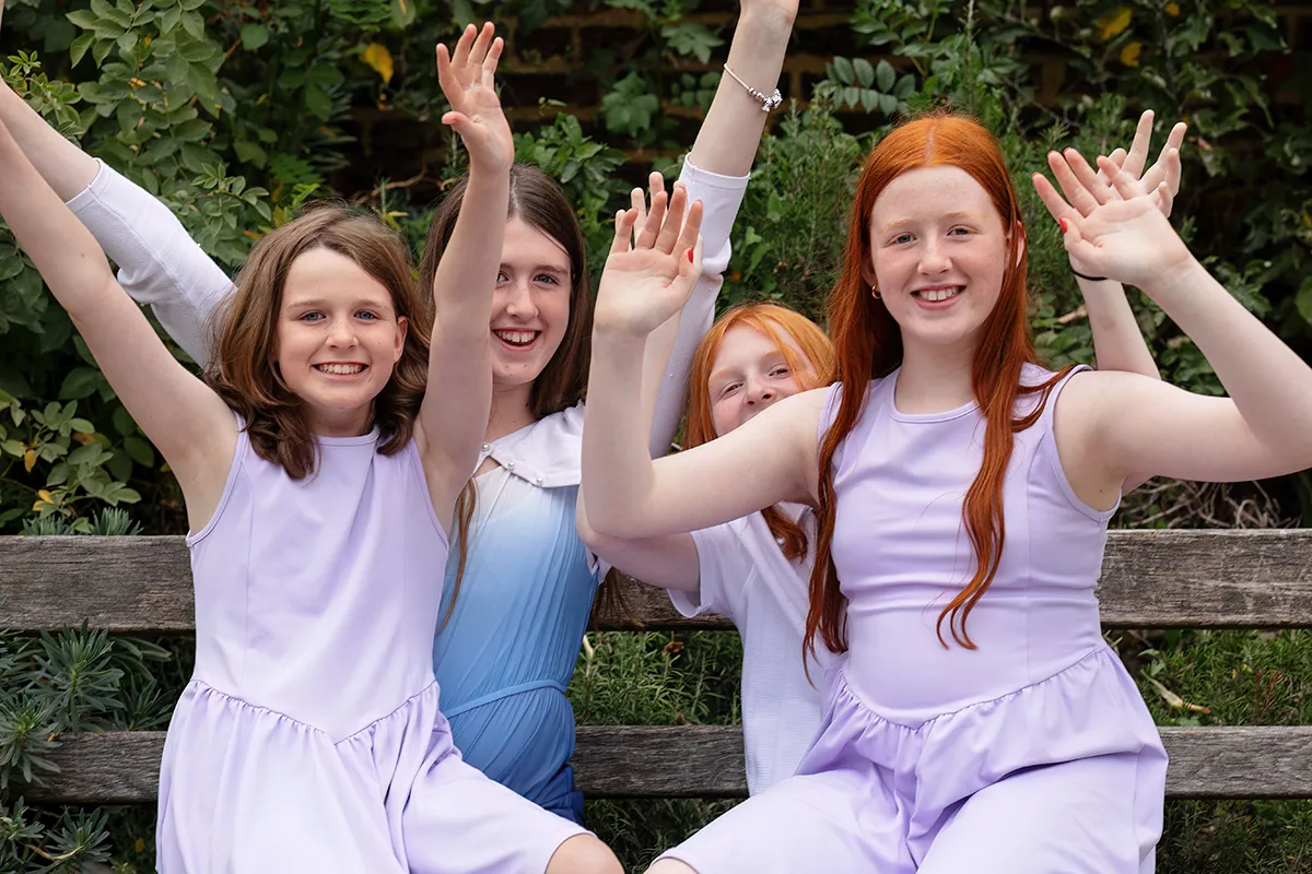 42_TJP_2025_08_16_0693 Four children with long hair, dressed in light-coloured clothes, sit on a bench outdoors at a Pitzhanger Manor Wedding Photography session, smiling and raising their arms amid lush green foliage.