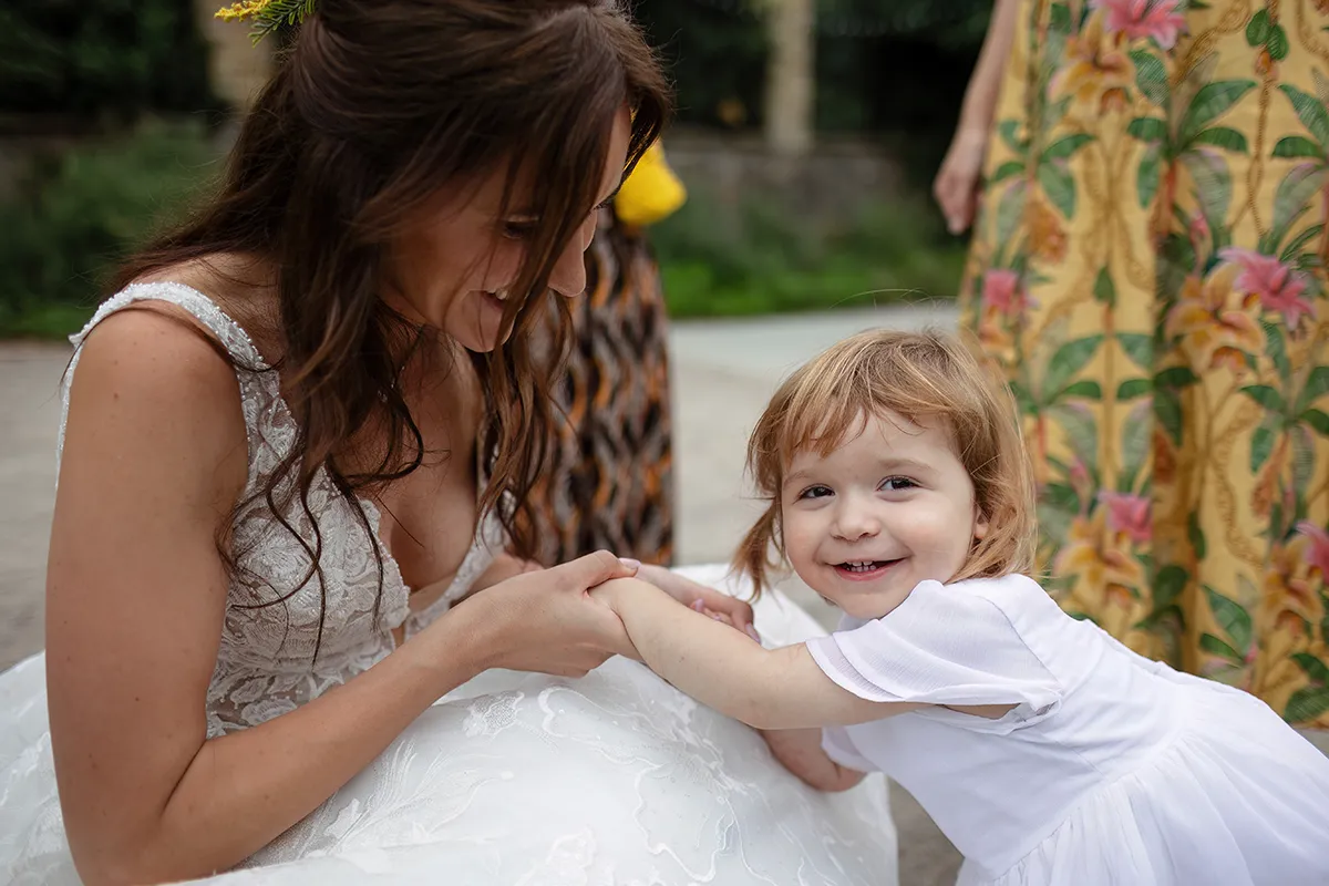 43_TJP_2025_08_16_0726 A smiling bride holds hands with a young girl in a white dress, both crouched outdoors. The girl looks at the camera whilst the bride gazes at her, surrounded by vibrant dresses—capturing a joyful moment of Pitzhanger Manor Wedding Photography.
