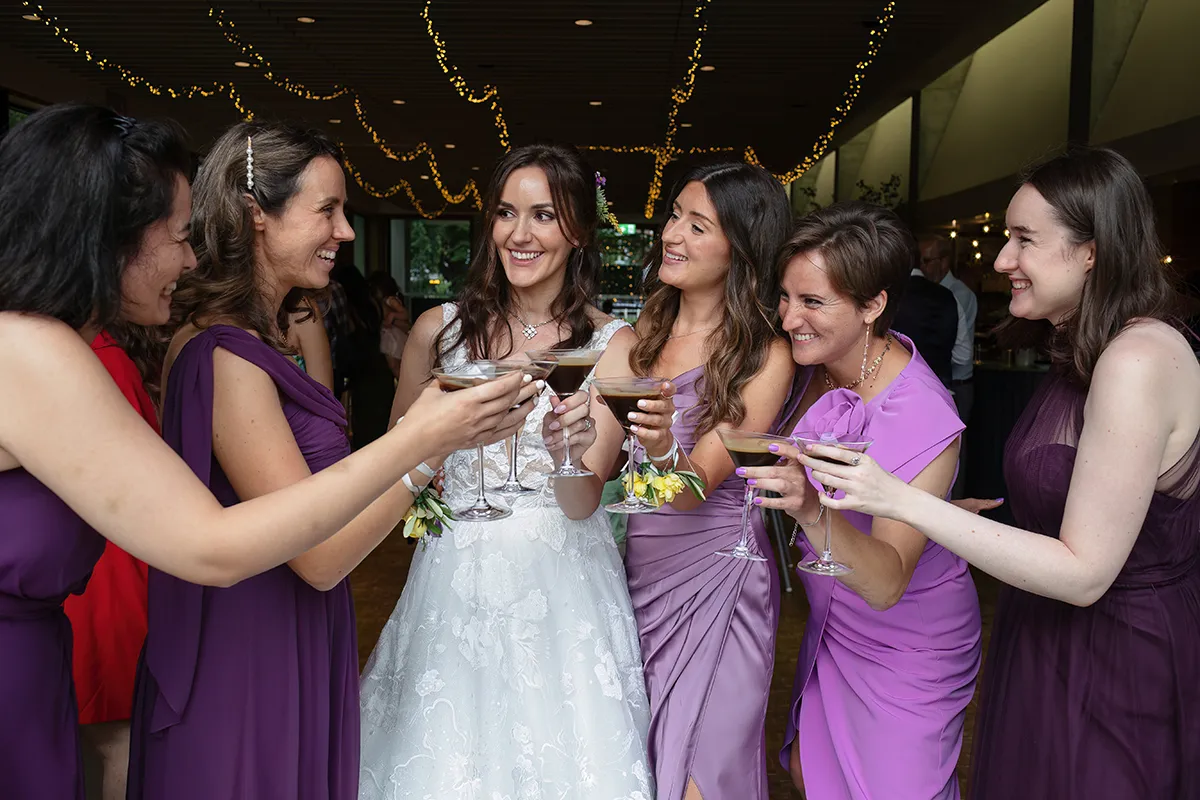 44_TJP_2025_08_16_0774 A bride in a white dress clinks cocktail glasses with five bridesmaids in purple dresses, all smiling and celebrating together indoors under fairy lights—capturing the joy of Pitzhanger Manor Wedding Photography.