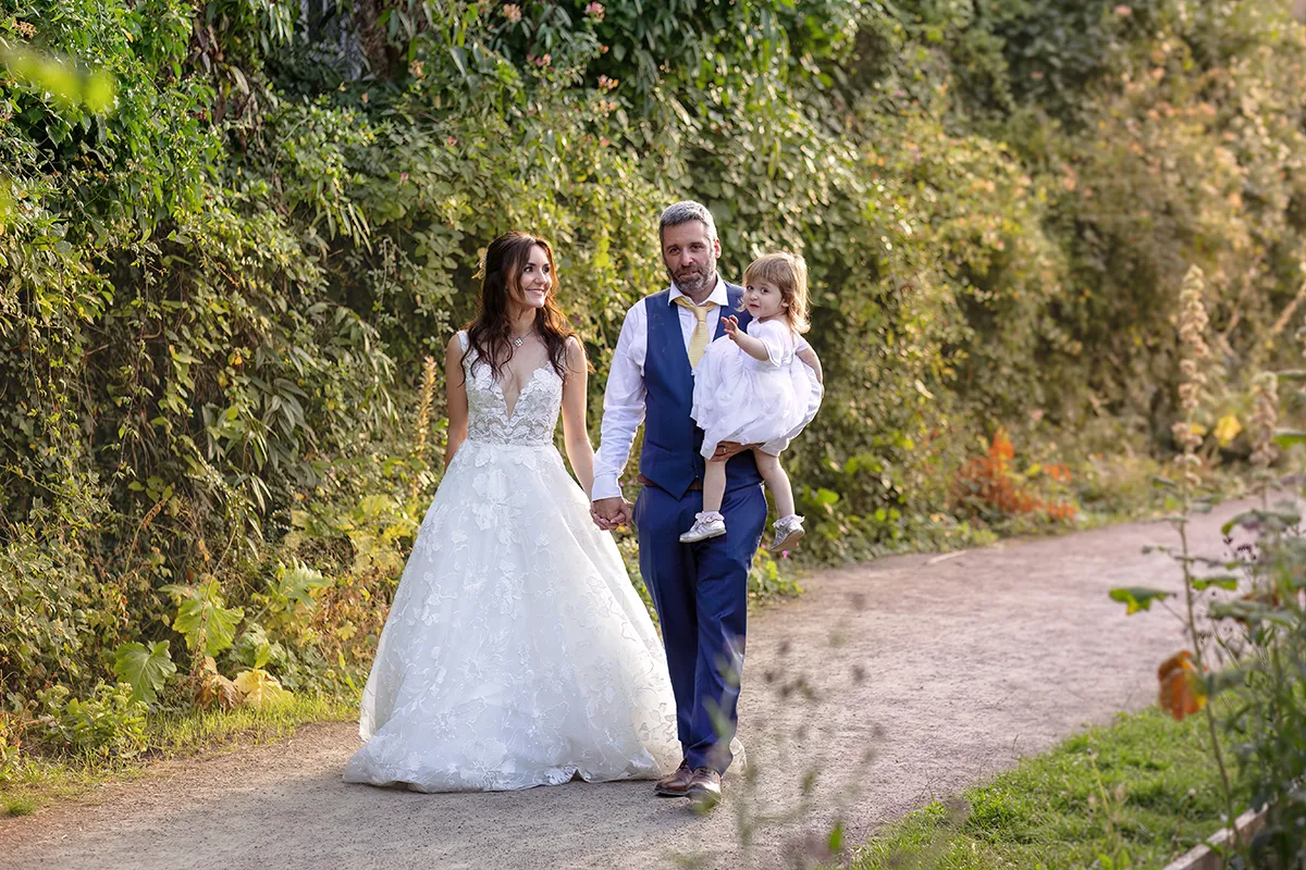 45_TJP_2025_08_16_0806 A bride in a white gown and a groom in a blue suit walk hand-in-hand on a path at Pitzhanger Manor. The groom carries a young child, and they are surrounded by greenery, capturing joyful Pitzhanger Manor wedding photography moments.