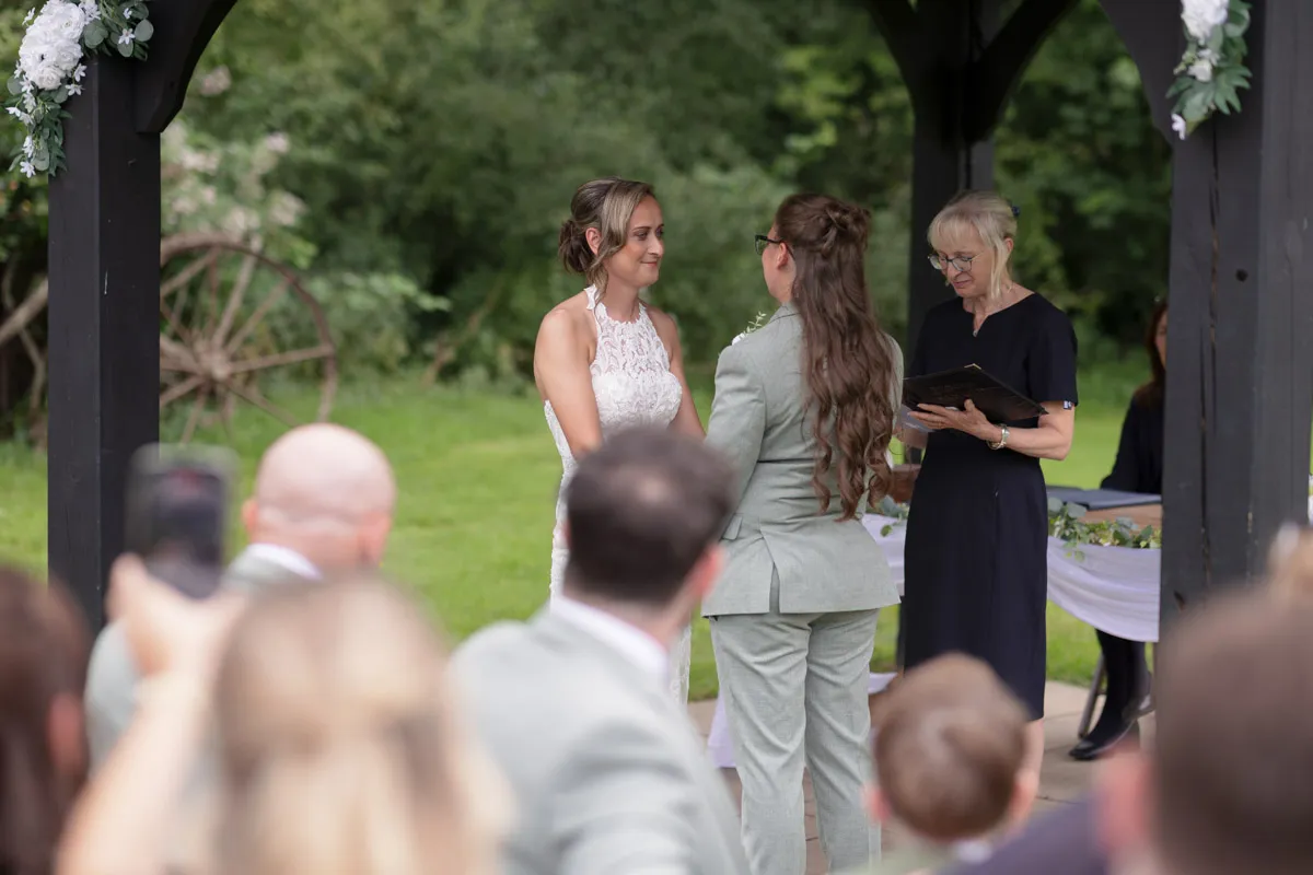 Two women stand facing each other holding hands at their Same-Sex Wedding at The Plough & Barn at Leigh, with an officiant reading from a book and guests watching. Lush greenery and rustic decor are visible in the background.