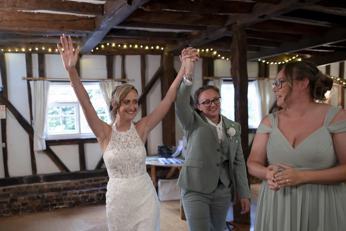 Two brides celebrate, holding hands; one in a white lace dress with arms raised, the other in a light suit smiling. A bridesmaid in a mint dress stands nearby inside the rustic venue with wooden beams and fairy lights, capturing the joy of their same-sex wedding at The Plough & Barn at Leigh.