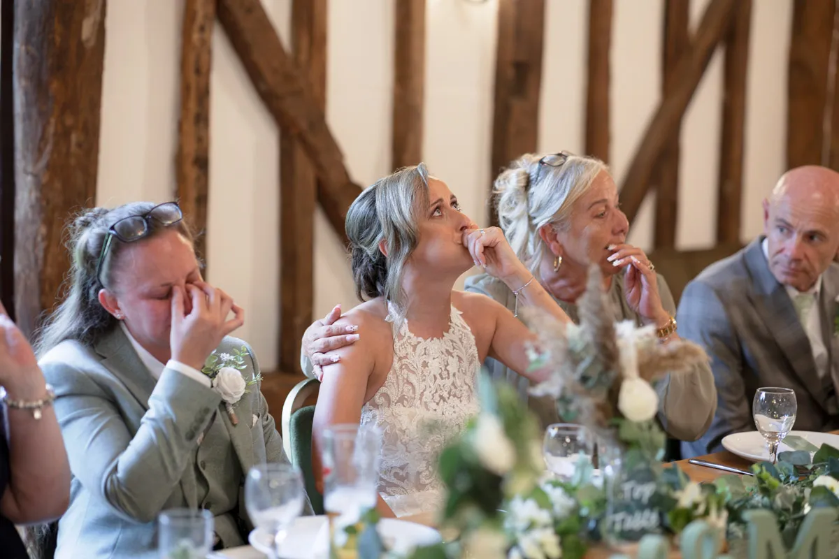 A bride in a white lace dress sits tearfully with two women and a man, all emotional, at a decorated table during a wedding reception in a rustic venue—capturing the heartfelt moments of a Same-Sex Wedding at The Plough & Barn at Leigh.