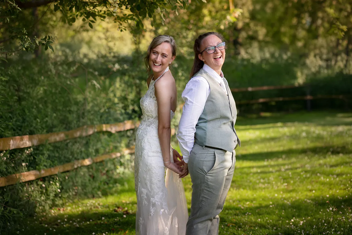 Two people in wedding attire stand back to back, smiling and holding hands outdoors during a same-sex wedding at The Plough & Barn at Leigh. One wears a white dress, the other a light grey suit, as sunlight filters through green trees.