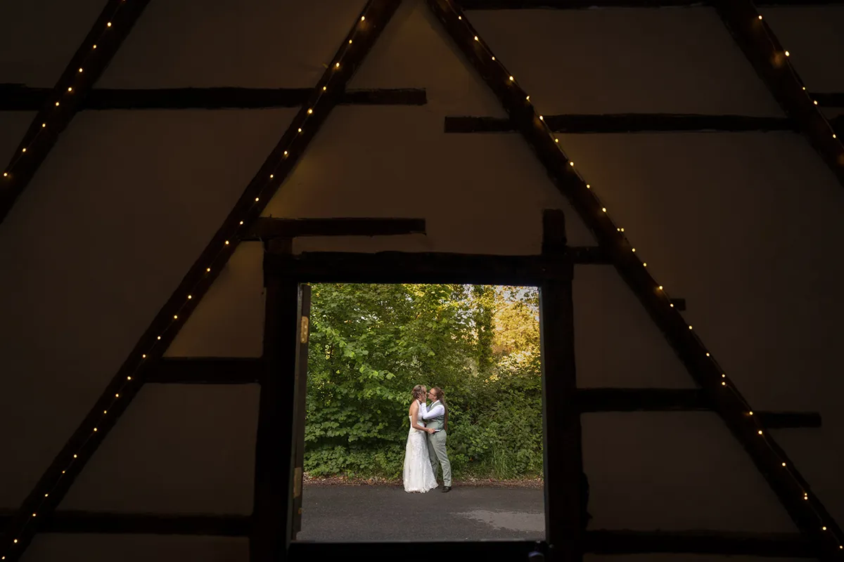 A bride and groom embrace outside, framed by a large triangular structure with fairy lights, viewed through a wide doorway and surrounded by greenery—a magical moment reminiscent of a Same-Sex Wedding at The Plough & Barn at Leigh.