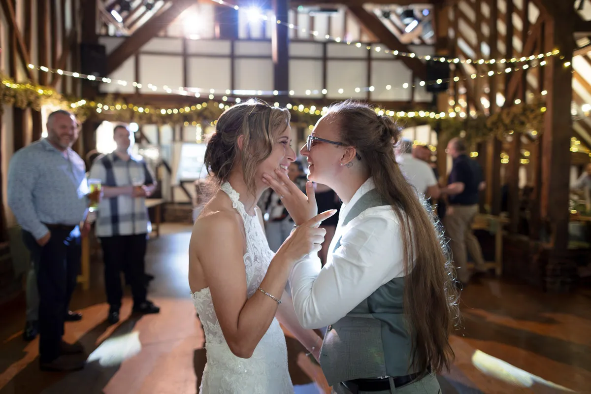 Two brides smile and laugh together whilst dancing at their Same-Sex Wedding at The Plough & Barn at Leigh, a rustic, wooden venue beautifully decorated with fairy lights and garlands, with guests celebrating in the background.